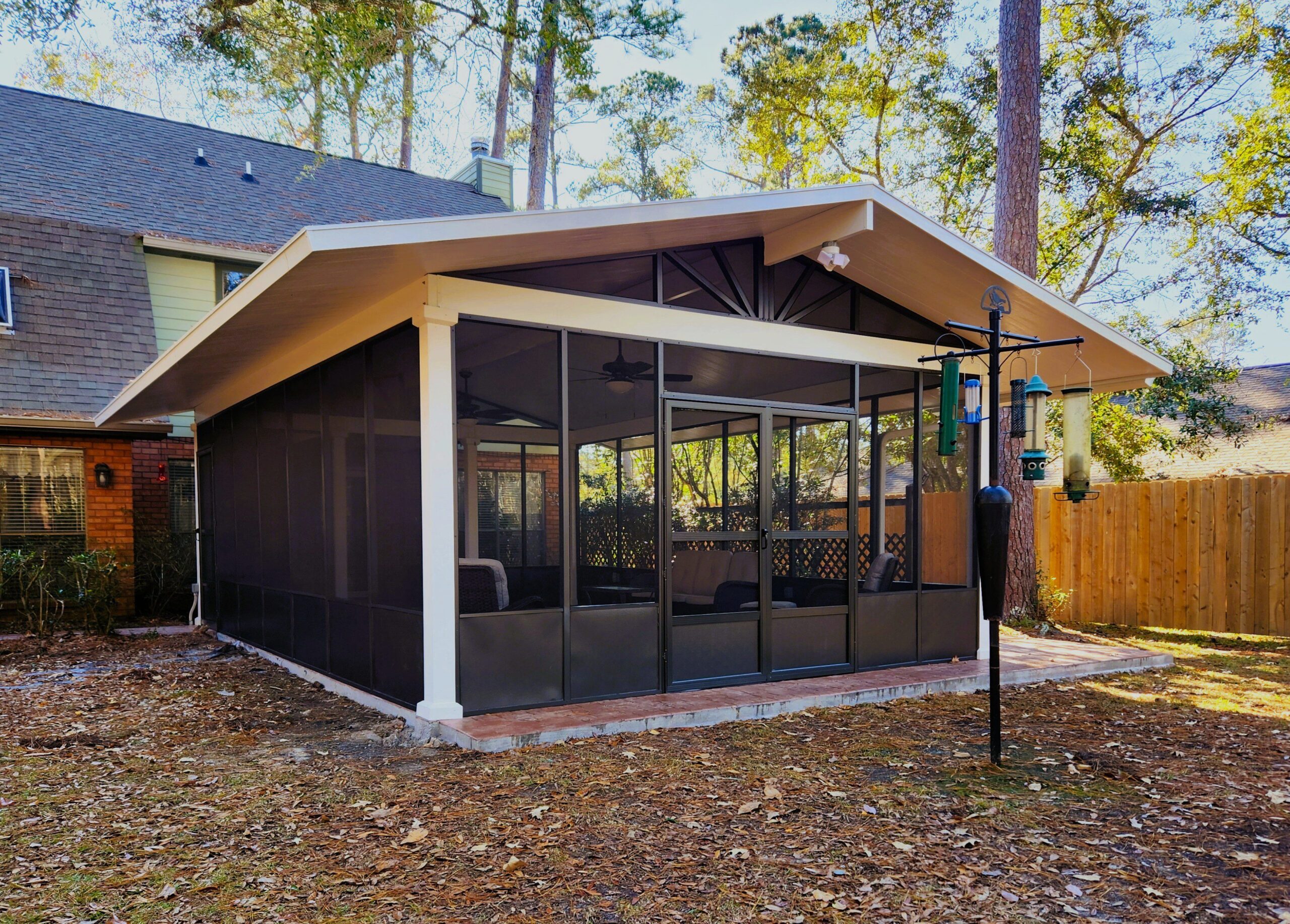 20241223_103729 - Capital Improvement A screened-in porch attached to a house, with a peaked roof and ceiling fans, is shown in a backyard with trees, a wooden fence, and bird feeders.